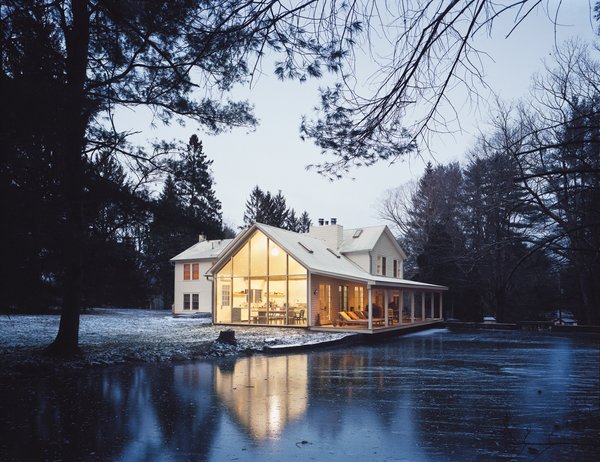 Hope Floats - Photo 5 of 12 - The Floating Farmhouse’s semitransparent addition has a roofline that matches the pitch of the original 1820s farmhouse. A porch, tucked under the side eaves, is cantilevered over a stream that runs through the property. Ikea loungers are illuminated from the interior by commercial gymnasium lights repurposed as pendant lamps.