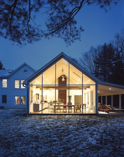 Hope Floats - Photo 8 of 12 - The Floating Farmhouse’s semitransparent addition has a roofline that matches the pitch of the original 1820s farmhouse. A porch, tucked under the side eaves, is cantilevered over a stream that runs through the property. Ikea loungers are illuminated from the interior by commercial gymnasium lights repurposed as pendant lamps.