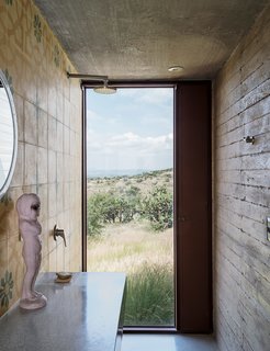 Mexican encaustic tiles with a geometric floral pattern from Mosaicos Terra line the bathroom in Austin's studio, where a full-height window near the wall-mounted shower provides a view to the expansive outdoor scenery. A vintage kewpie doll sculpture sits atop the custom terrazzo-and-granite counter; the steel mirror is from Artes de Mexico.