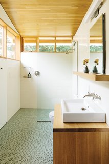 In the bathroom, a thin pane of glass separates the shower; an Aquaplane sink by Lacava hovers above <br><br>a built-in vanity illuminated by a lean Adelphi light by Oxygen Lighting; and blue-green glass penny tiles by Terra Verre decorate the floor. The absence of a door, combined with windows on two sides, makes the bathroom feel like a continuation of the overall space.