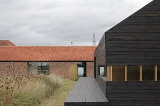 A Pair of English Barns Hide Unabashedly Bold and Budget-Friendly Minimalist Interiors - Photo 5 of 14 - For a bit of elevation in the overwhelmingly horizontal compound, step onto the deck of the Stealth Barn. A strip of mowed grass delineates a path between the two structures; otherwise the grasses grow wild.