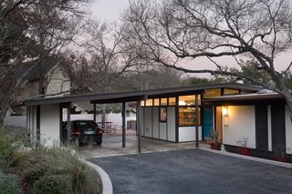 A wide view of the renovated home situates it as it sits on Austin soil. Where the gentle swoop of the driveway meets the overhanging garage, the home's patio is just visible. A light in the new kitchen window further integrates the home with the neighborhood just beyond it.