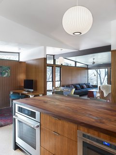 A post-renovation view of the kitchen shows it opening into the family room. Replacing the former white cabinets are Island drawer fronts and wall panelling of teak wood and reclaimed American elm countertop, milled by Vintage Material Supply. The differing grains of the teak veneers and elm countertop vadd complexity and rhythm to the kitchen's wood motif. Stuc Pierre plaster ceiling selected by the homeowner, Sloan Houser, adds an airy feel to the opened space.