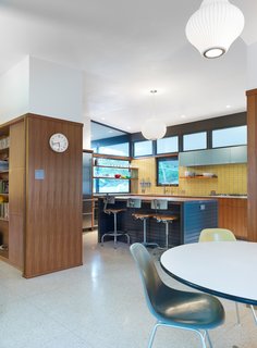 The open dining area and kitchen of a midcentury renovation in Austin shows the existing terrazzo flooring, the plaster ceiling and the kitchen's tile backsplash, Fireclay Debris series in ‘Daffodil,’ working in harmony. Floating shelves in the kitchen window and a stainless steel countertop surrounding the kitchen's burners rest just beneath the backsplash. A Boca Raton Blue upper cabinet and Nelson pendants add interest and dimension to the kitchen's warm combination of wood, yellow and blue. Vintage chairs, seated at the kitchen island and in the front right corner at the dining table complete the design scheme.