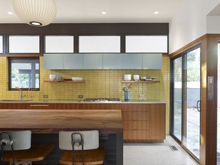 A view of the kitchen's back wall shows the bright marriage of the yellow Fireclay tile backsplash with incoming light from the adjacent sliding door and the row of windows just above the hanging Boca Raton blue cabinets. Basked in light, the new kitchen displays an organized and cheerful aesthetic.