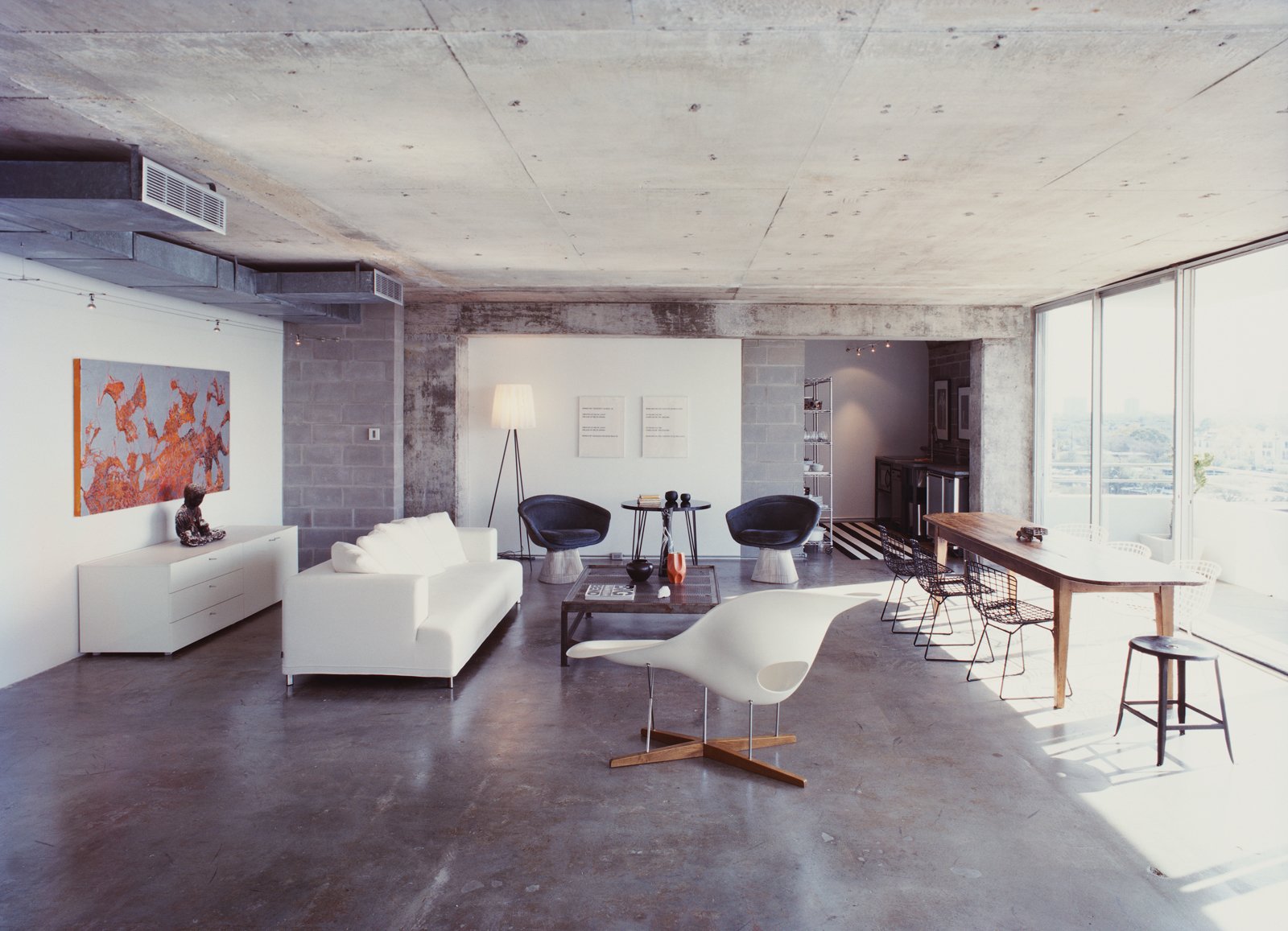 A Philippe Starck standing lamp and an Eames chaise longue bracket the living room; two Lawrence Weiner prints hang behind a pair of Warren Platner chairs and a table purchased from a River Oaks estate sale; at far left of the room, a partial wall of new cinderblocks hides a return air vent.