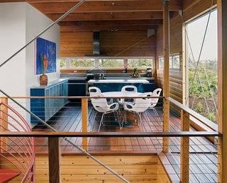 The home's upper floor features copious amounts of untreated pine. Five large cross-braces keep the timber frame rigid. The royal blue Ikea kitchen, Noguchi Cyclone dining table for Knoll, and Karim Rashid Oh chairs for Umbra all made the trip from the mainland in a twenty-foot-long shipping container. Above the wall-mounted credenza, a colorful painting of the Puma moonrise is by local artist Arthur Johnson.