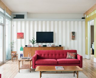 The Shipping Muse - Photo 21 of 23 - The couple had a big flat-screen TV but didn't want it to be the focus of the living room. Placed on the wall behind one of the red couches, it hangs quietly without drawing attention to itself but is in perfect position for TV-watching from the second couch (not pictured). Below the television is a console that Feldmann's mother picked up at a thrift store for $50. Feldmann had her reservations about the piece at first but after they took off its original base and lifted it on two-by-fours for an elevated look, she was sold.