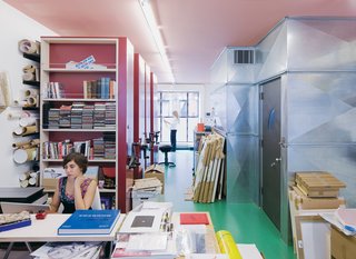 The couple’s archival photographer Alyssa Gorelick sits in front of Lawrence’s sliding red storage units while Alice reviews some papers in the background.