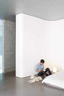 The curving white wall in Atherton’s bedroom is optimally sited to capture shadows from the redbud tree outside his window. Pip, the dog, will have to content himself with concrete floors—–at least until his housemates buy a couch. Or a rug.