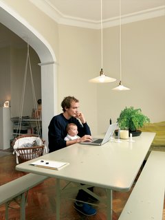 Kind of New - Photo 5 of 15 - Thomas Lommee and baby Emilia Luz work at the beer-garden table under hanging lamps given to the couple by friends. The baby bassinet, which appears to be made from a wicker laundry basket and some sturdy rope, was also a gift from friends.