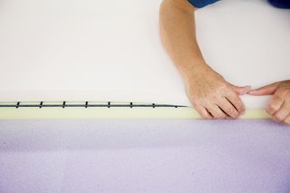 Ruché Sofa - Photo 6 of 11 - A technician sprays a sheet of pliable purple memory foam with a water-based adhesive and then carefully folds it over the other two foam layers and a steel spring grill to complete the backrest.