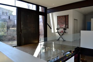 The house's main door is a pivoting thousand-pound piece of steel that opens with a push of a finger. You can see the recycled waste-concrete retaining wall through the glass, with Grandmother's Cabin visible at the top of the lot. Glass guardrail panels in the foreground sit on the steel floor beam, which also serves as an ad-hoc message board with magnetic letters. Photo by J.C. Schmeil.