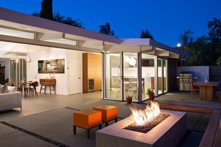 In the outdoor living area, orange Primary Pouf stools by Quinze & Milan and an ipe bench surround the central fire pit. An outdoor kitchen neighbors its interior counterpart. In addition to a grill, it accommodates a table and bench by Kayu.