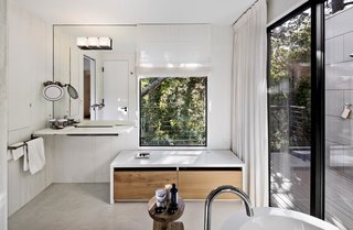 The updated master bathroom features white concrete floors, painted wood walls, and veneer plaster ceilings. Light streams in through the original window, and an added sliding door opens to an outdoor shower. A single Washplane sink by Omvivo sits beneath the mirror, with additional faucets for the shower and Duravit bathtub by Hansgrohe. A stool from Roost holds bath essentials.