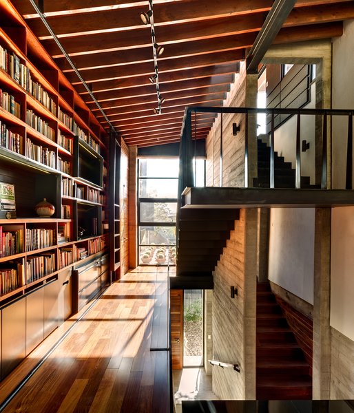Suspended over the living room, the library bridge connects the second floor rooms of the house. The transitional space contains built-ins and desks where the owners and their three-year-old daughter can sit and read. The steel of the staircase is supported entirely by the adjacent concrete wall. A marriage of concrete, wood, and glass is used in the space, and the large windows let light in and offer a view of the roof garden. Photo 8 of Hybrid Home modern home