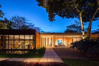 With library's Cor-Ten shield open, the home’s facade is seen at night. The L shaped pedestrian pathway forces visitors to travel around the perimeter before entry, gradually experiencing the architectural composition step by step.