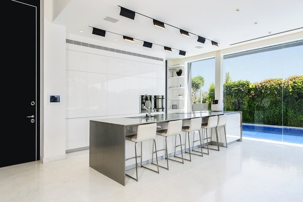 A well-lit and open ground floor with a strong interior-exterior connection was at the top of the family’s wish list. The kitchen makes the most of a compact space through its intimate relationship with the adjacent pool and public outdoor space. The spaces features hardware-free white cabinets and a stainless steel center island. The white kitchen floors provide visual continuity with the white pavement directly outside. Photo 3 of Rishon Le Zion 3 modern home