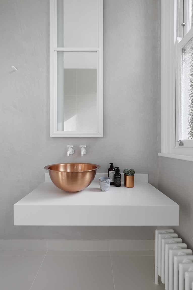 Metal details in the home transition to gradually darker tones as the spaces become increasingly private. In the guest bathroom, a solid brass bowl sink rests on top of a custom Corian base. White Dornbracht wall-mounted faucets sit below a custom mirror cabinet. In the bathroom and throughout the home, interior accents by Gunter & Co make elegant finishing touches.
