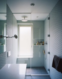 Triple-glazed, frosted windows emit soft, filtered light against pale gray and blue surfaces inthe master bathroom. Lucian Field matte-glass and Lucian Mosaics penny round tiles, both by Ann Sacks, line the floor and walls.