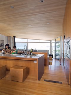 Lam and her son Max prepare a snack together in the open-plan kitchen at the center of the house. The fir floor that covers the living space steps downward twice, creating a grade change that roughly follows the topology of the site. The ceiling is sheathed in untreated hemlock; the custom stools were designed by Brent Comber. 
