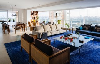 Overlooking a Niemeyer Masterpiece, This Flat is Filled with Brazilian Modern Gems - Photo 4 of 9 - Furniture groupings and bright, dramatic accents—like the sapphire banana-fiber rug by Kamy Maison in the main living area—further divide the interior into distinct yet flexible stations. The trio of seating options includes wood-and-cane Cosme Velho armchairs by Claudia Moreira Salles, a plush Soft Dream leather sofa by Antonio Citterio for Flexform, and a blue Fergana sofa by Patricia Urquiola for Moroso. Just beyond, a Fina table, also by Salles, joins Marta chairs by Aristeu Pires and Torch pendants by Sylvain Willenz to form the dining area.
