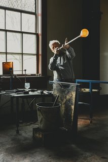 Master Glassmaker Simon Pearce's Sustainable Factory in Vermont - Photo 3 of 16 - Designer Simon Pearce, shown at his factory in Windsor, Vermont, makes handblown glass the same way it’s been made for thousands of years, by melting the raw ingredients in huge ovens, then gathering the molten material onto iron blowpipes. A finished Westport footed glass sits on a nearby workstation as a point of reference. 