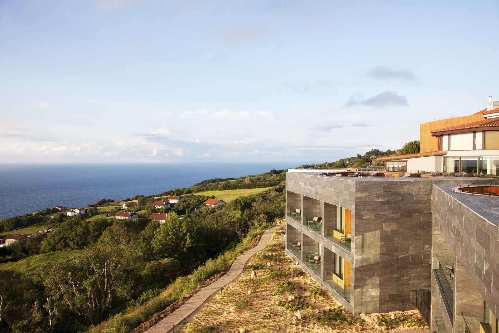 Comprised of five stone cubicles that are built into the hillside facing the sea, the structure features a rooftop deck with slightly sunken outdoor pits for guests to enjoy. The terraces are connected by a series of planked decking and landscaped gardens that offer a spectacular panoramic view of the coastline.