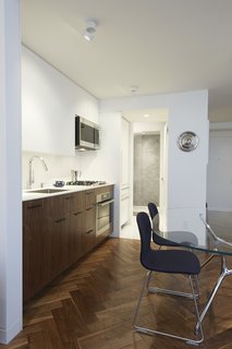 The galley kitchen features a slim Corian countertop and backsplash over rich walnut cabinetry. In the pantry and bathroom areas beyond, white terrazzo flooring replaces wood. The wall at the back of the shower is clad in large-format Blue de Savoie stone tiles.