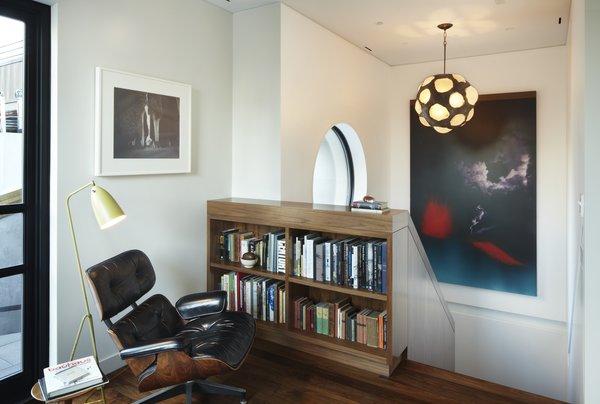 The upper landing in the White Street Residence provides a pausing point when going to and from the roof garden. A walnut bookshelf, an extension of the walnut knee wall below stores a collection of the owner's art books. The large circular window which overlooks the roof garden can be seen in the background. A vintage Grasshopper floor lamp by Greta Grossman provides reading light while relaxing in the Eames 670 lounge chair. Photo 12 of White Street Residence modern home