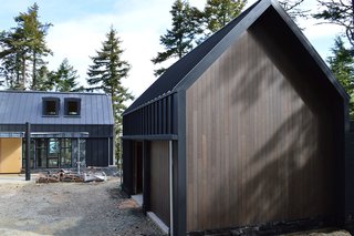 The garage exhibits the same black steel cladding and stained cedar siding as the main structure. Flat dormer windows on the house provide natural light and add visual interest to the overall shape.