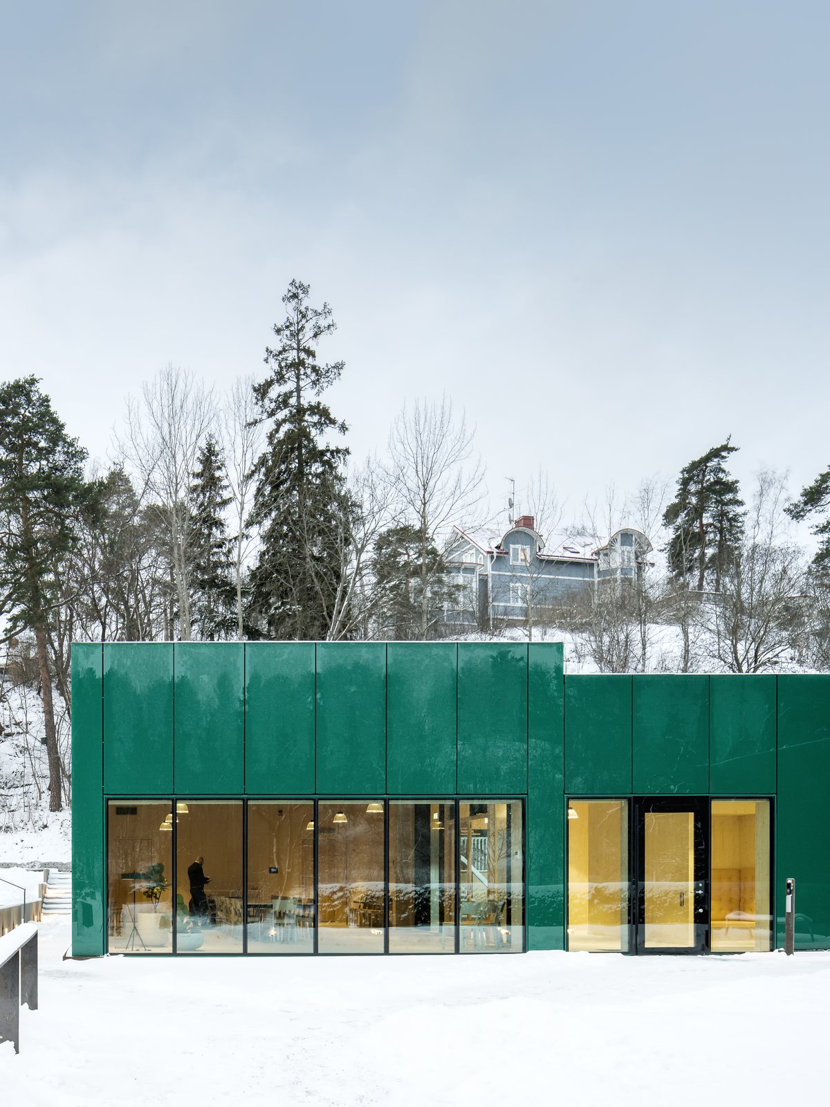 The bold, emerald hue complements the cemetery's lush surrounding gardens. The green glass reflects the surrounding vegetation, integrating the building into the garden spaces framed by the cemetery’s hedges.