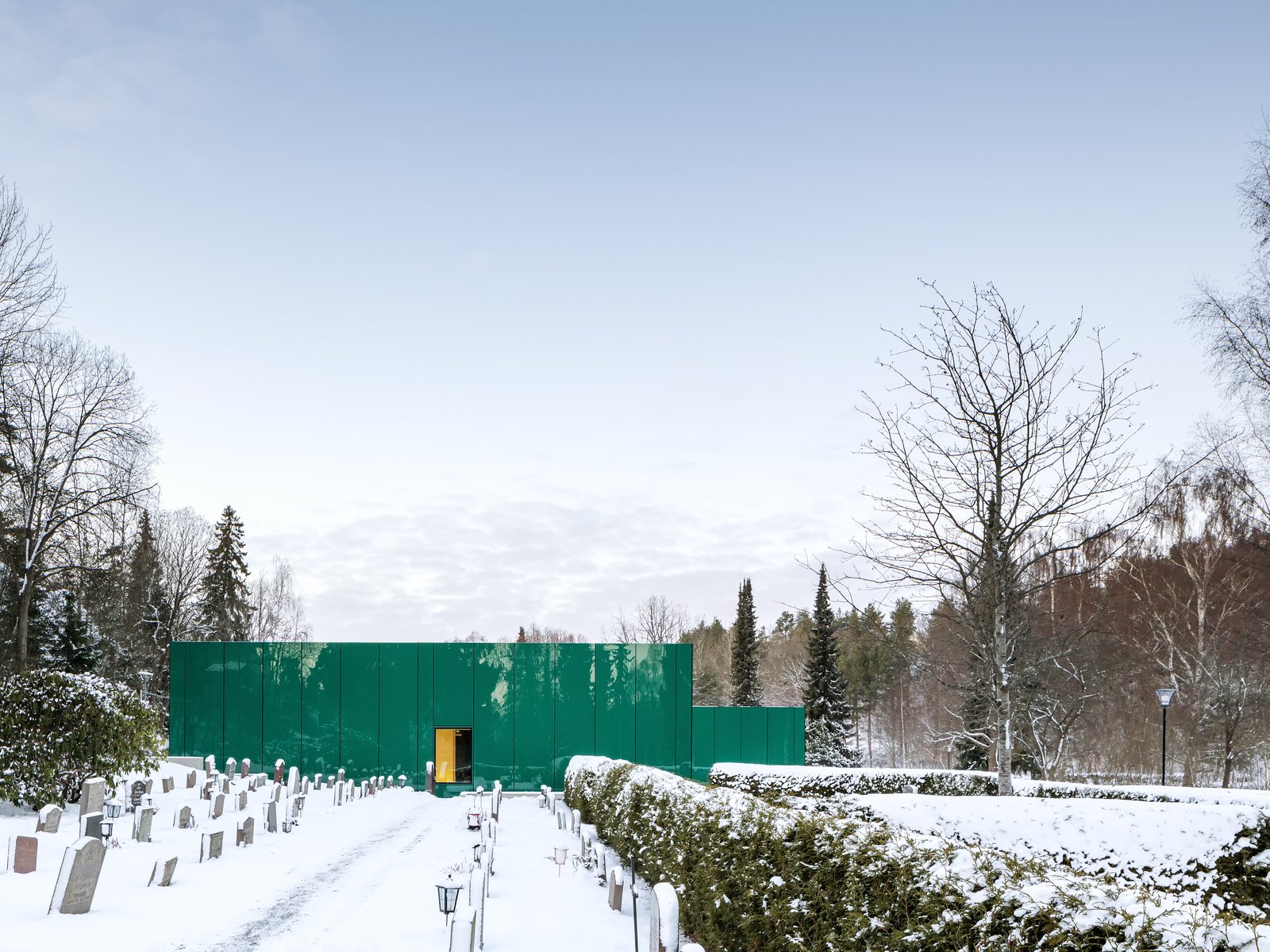The green glass reflects the surrounding vegetation, integrating the building into the garden spaces framed by the cemetery’s hedges.