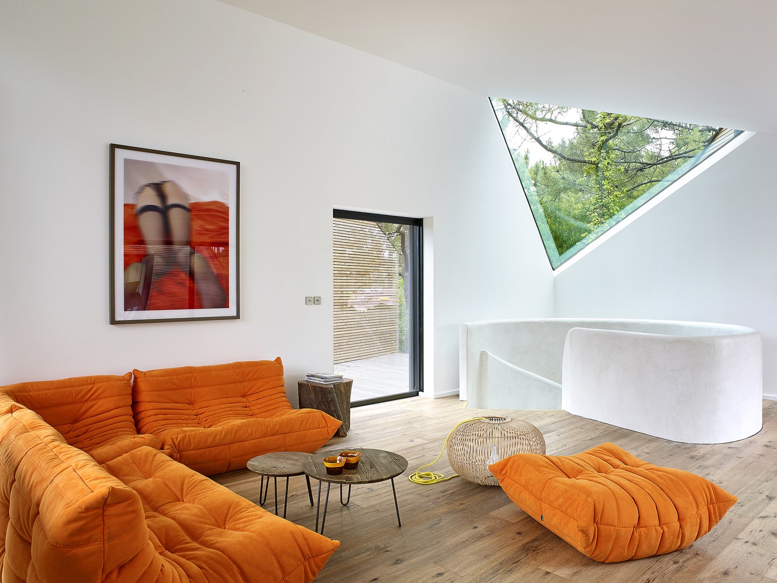 An orange Ligne Roset Sofa provides a comfortable space for rest and relaxation. A triangular window provides a picture of the tree canopy beyond, while drawing in natural light.