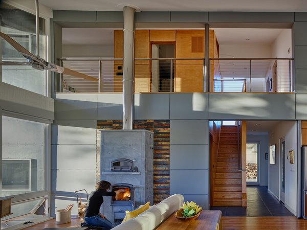 Modern home with living room, sofa, coffee tables, two-sided fireplace, medium hardwood floor, and wood burning fireplace. Heating is primarily provided by the dual-sided Tulikivi stove. The Owner has been experimenting with cooking chicken, pizza and bread in the upper oven. The cementitious board walls (blue) run from exterior to interior, creating a series of thresholds that demarcate the public and private realms of the house. Photo 3 of Mill Pond House