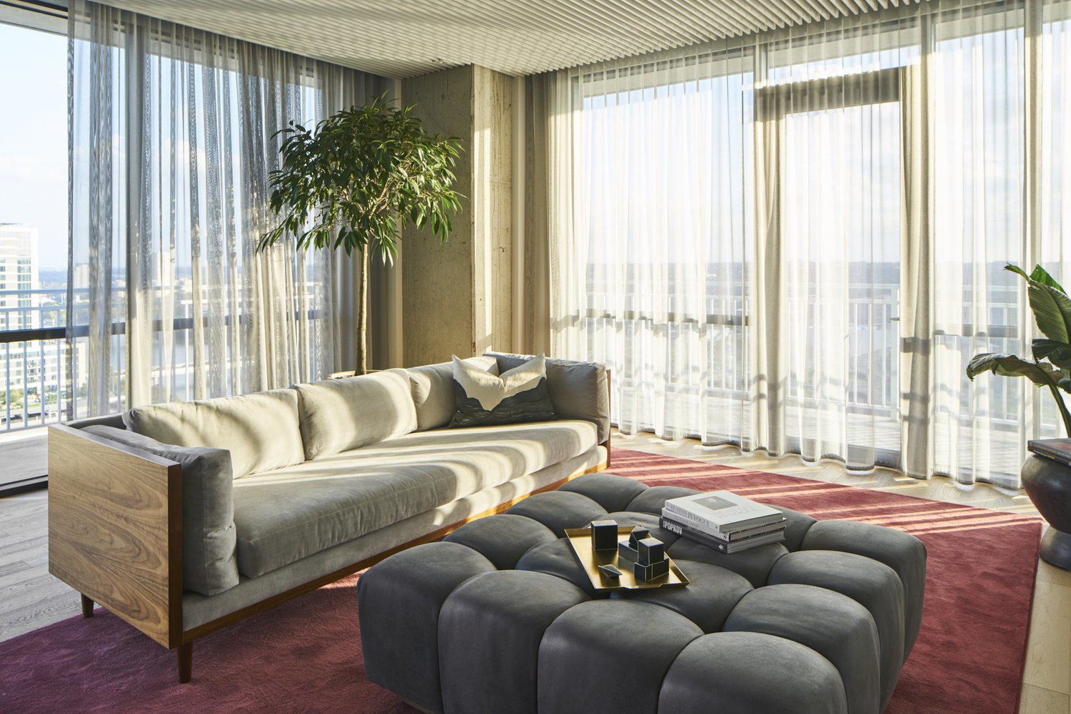 Living Room with wraparound balcony, cement walls, bubble tufted ottoman, wooden couch base, sheer curtains and red area rug.